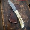 Custom handmade 7-inch Damascus folding knife with bone handle, brass bolster, and engraved name “Keuck,” forged by Damascus Edges, displayed on rustic wooden bench with leather sheath.