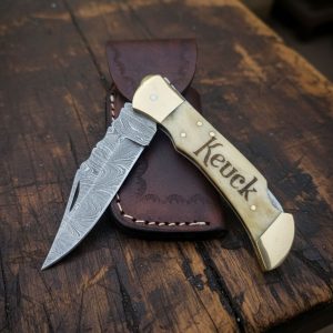 Custom handmade 7-inch Damascus folding knife with bone handle, brass bolster, and engraved name “Keuck,” forged by Damascus Edges, displayed on rustic wooden bench with leather sheath.