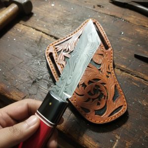 Hand-forged Damascus tanto knife with red wooden handle and hand-tooled leather sheath on a rustic American workbench
