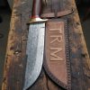 Hand-forged 13-inch Raindrop Damascus Bowie knife with polished wooden handle and engraved leather sheath labeled TRM, crafted by Damascus Edges on a rustic American workbench.