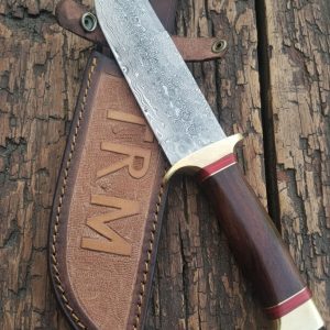 Hand-forged 13-inch Raindrop Damascus Bowie knife with polished wooden handle and engraved leather sheath labeled TRM, crafted by Damascus Edges on a rustic American workbench.